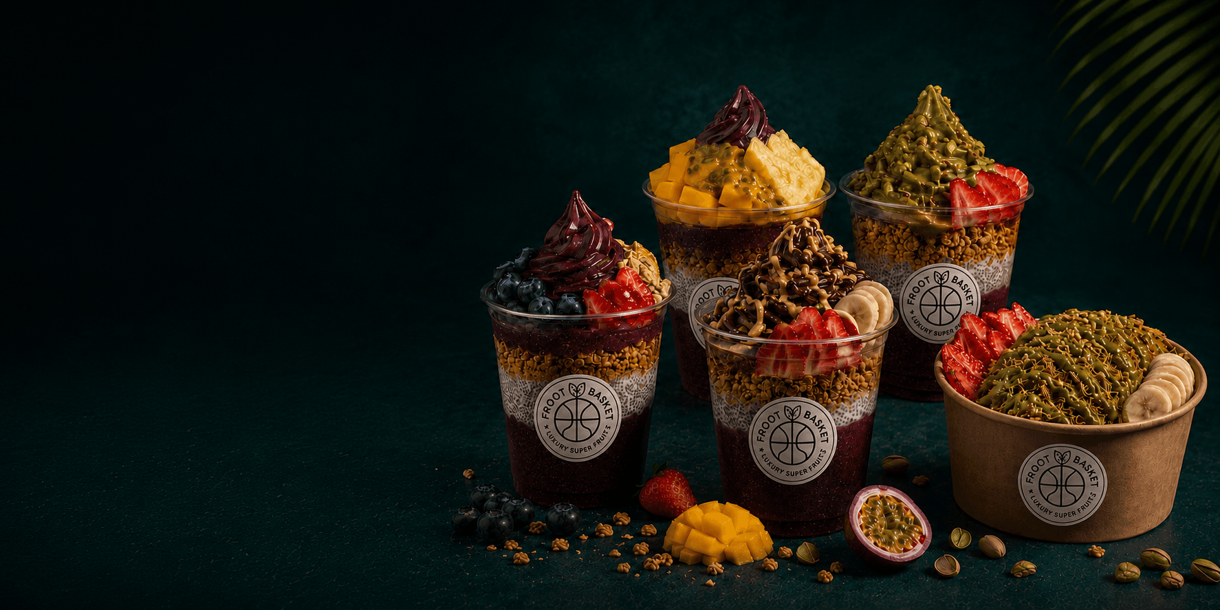 Vier Froot Basket Açaí Becher und eine Açaí Schale mit Früchten, Granola und Pistazien-Topping vor dunkelgrünem Hintergrund mit Palmwedel